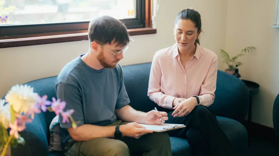 A man writes on a clipboard while a woman in a pink blouse sits beside him, engaged in conversation