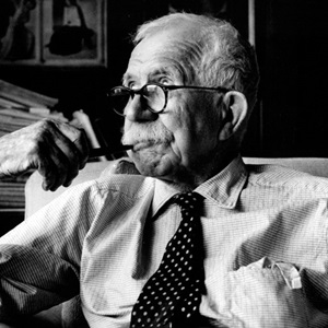 Sir Walter Murdoch as an eldery man, sitting in chair while smoking an old fashioned pipe.