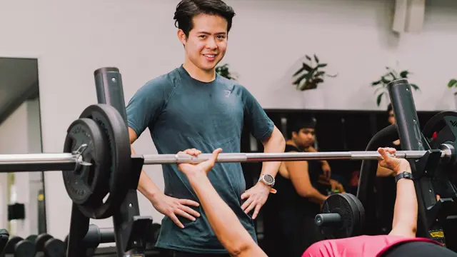 Allied health student assisting a client with a bench press in a gym