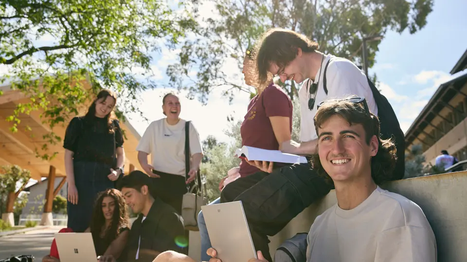 A group of students relax on the grass beneath large trees on a sunlit university campus.