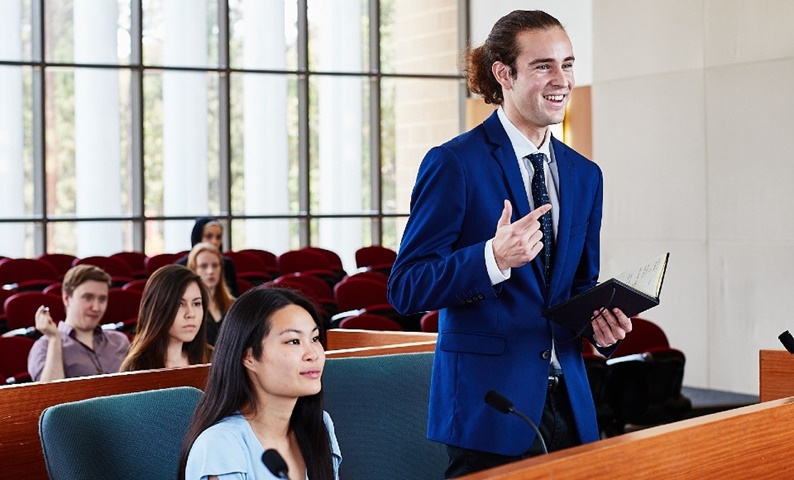 Male law student standing in Moot Court. A female law student is sitting beside him, with others in the gallery behind.