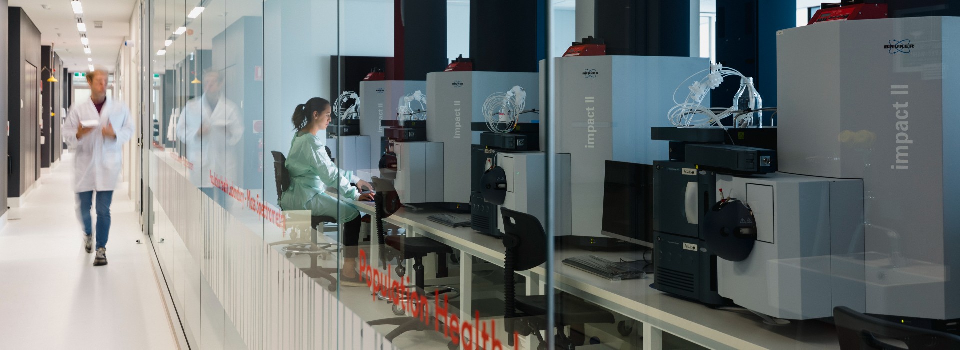 A researcher working in the Australian National Phenome Centre's Population Health Laboratory while another researcher walks past in the hallway outside.