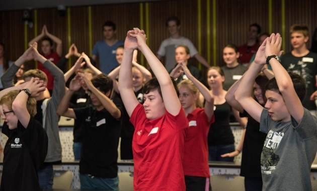 High school students in a lecture theatre, standing and clapping their hands above their heads.