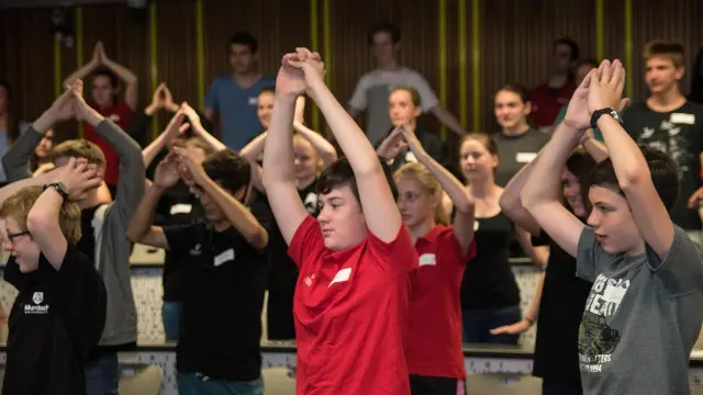 High school students in a lecture theatre, standing and clapping their hands above their heads.