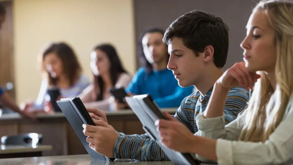 High school students sitting at desks reading on tablets.