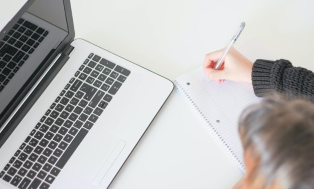 A brown-haired person sits in front of a laptop computer and makes notes on a notepad.