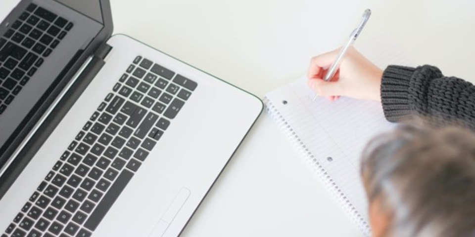 A brown-haired person sits in front of a laptop computer and makes notes on a notepad.