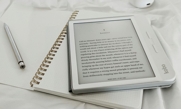 An e-reader rests on top of an open notebook, next to a silver pen.