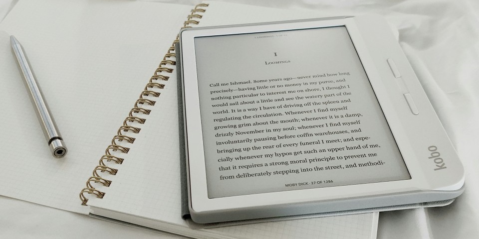 An e-reader rests on top of an open notebook, next to a silver pen.