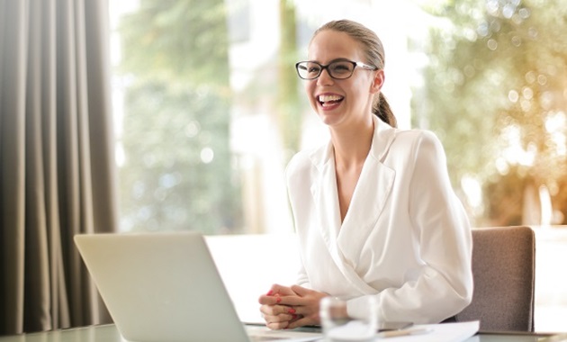 Woman in white blouse and glasses sits at a laptop, smiling.