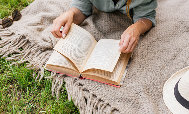 A woman relaxes on a picnic blanket on the grass, reading a book with had and sunglasses next to her.