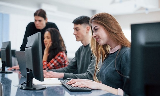 Three students work at desktop computers while a trainer assists them.