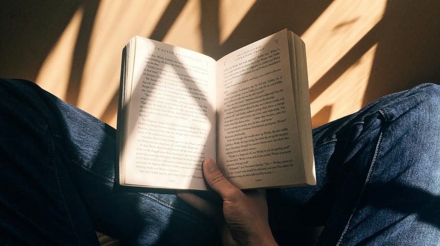 A person sits cross-legged in dappled shade reading a fiction book.