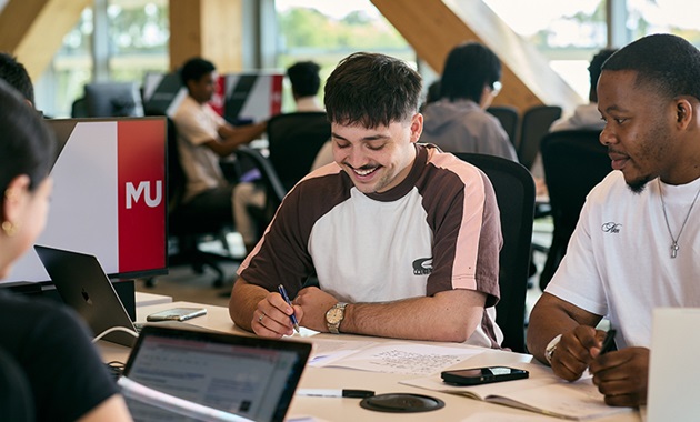 A group of students sitting at a table. One student is writing on page and smiling, and another student looks over their work