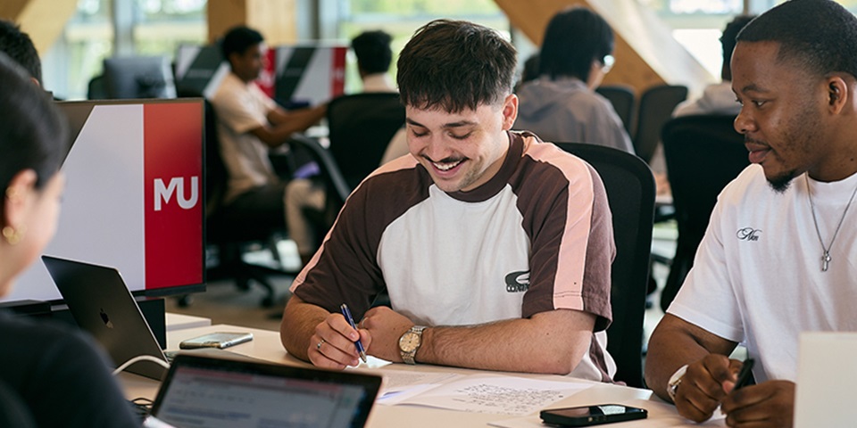 A group of students sitting at a table. One student is writing on page and smiling, and another student looks over their work