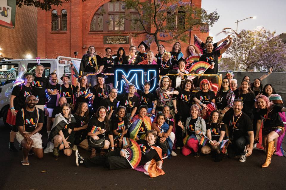 About 50 Murdoch staff and students, all smiling in Pride-themed Murdoch shirts, stand before a float with a glowing neon logo