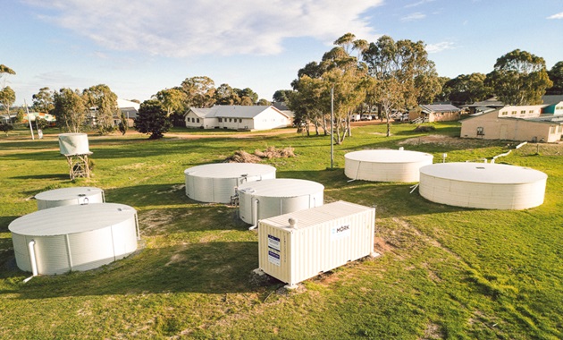 A white shipping containing in a field in front of a school surrounded by water tanks.