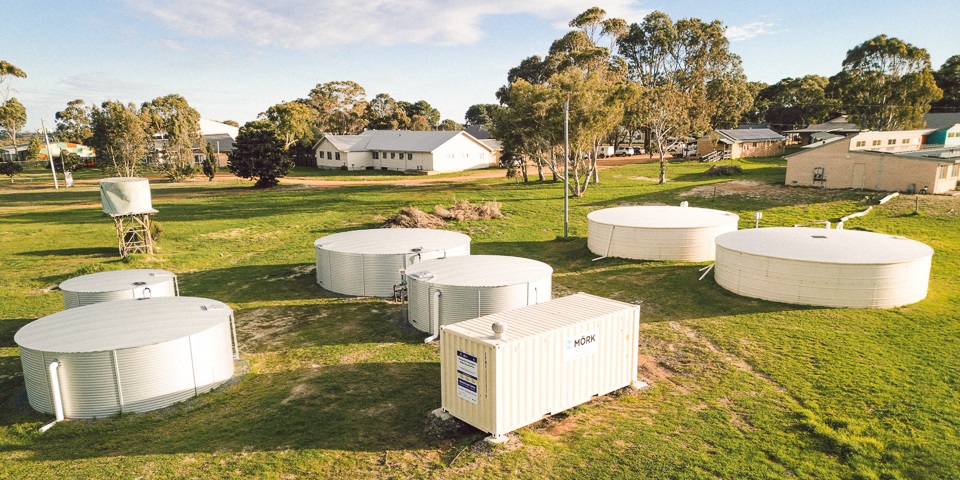 A white shipping containing in a field in front of a school surrounded by water tanks.