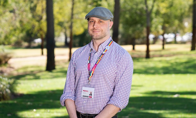 Man in check shirt and cap standing in a park looks towards camera