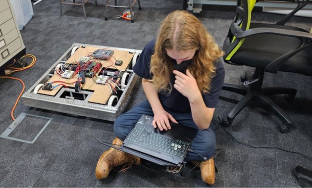 A person sits on the floor with a laptop, robotics equipment beside them