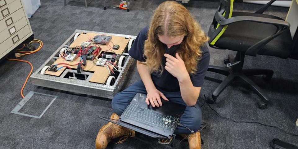 A person sits on the floor with a laptop, robotics equipment beside them