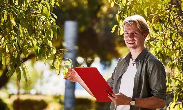 Student stands in bushland inspecting a bush with a clipboard