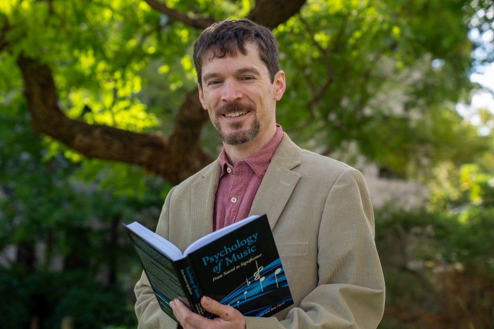 Associate Professor Jon Prince standing under a tree holding a book.