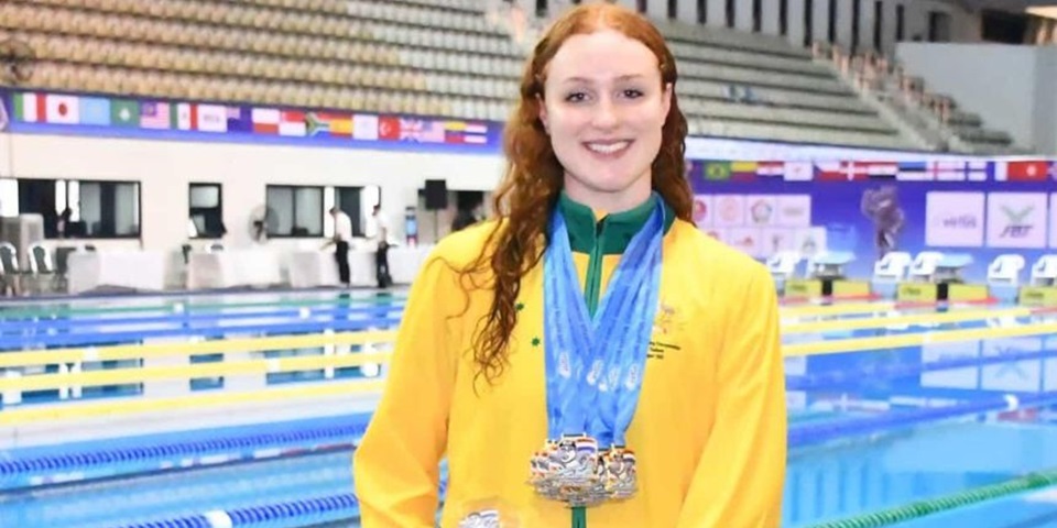 Murdoch University Elite Athlete Kate Wallington at the 2025 Virtus World Swimming Championships in Bangkok with medals around her neck and holding a trophy. She's standing in front of a swimming pool that is split into lanes for racing.
