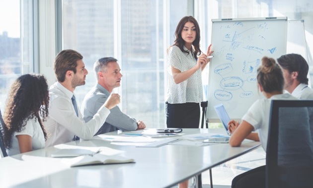 Lady speaking to a group of colleagues in front of a drawing board