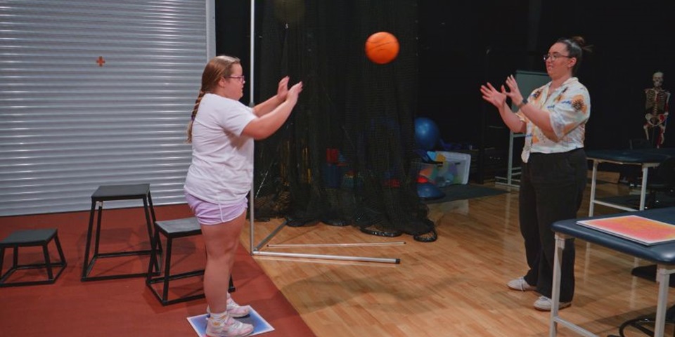 Participant Poppy and facilitator stand in the gym, passing a basketball to one another.