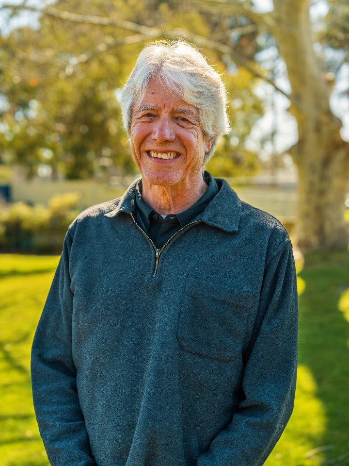 A headshot of Professor Wendell Ela standing in a blue jumper in front of a tree.