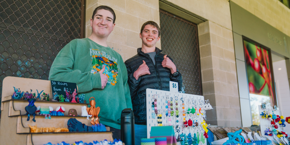 Two young men stand behind a market stall with colourful handmade craft goods on the table