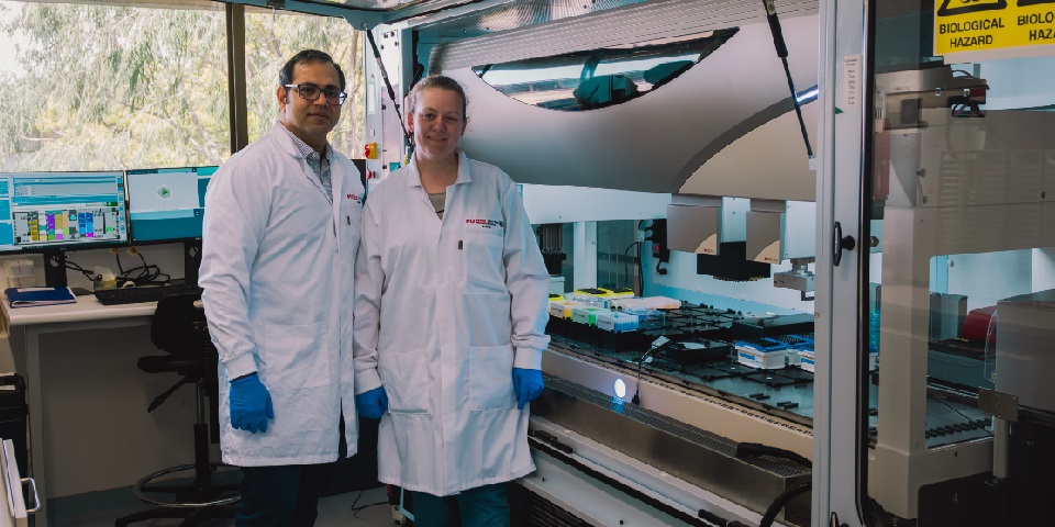 Two scientists stand in front of automated sample processing hood