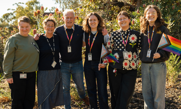 Six people standing in front of banksia trees smiling and holding progress pride flags.