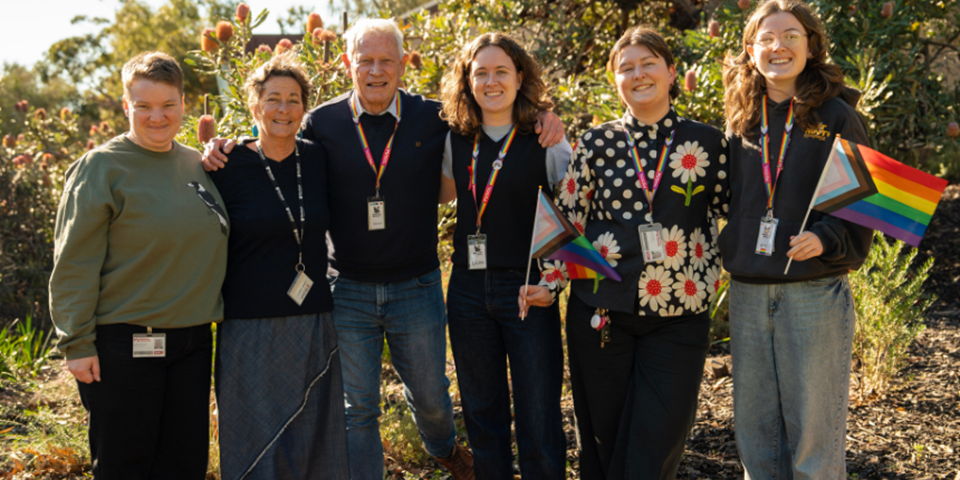 Six people standing in front of banksia trees smiling and holding progress pride flags.